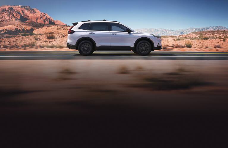 2026 Honda CR-V Sport Touring Hybrid in Platinum White Pearl profile driving on a scenic desert road (Compact Crossover SUV).