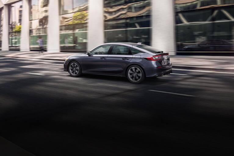 2027 Honda Civic Hatchback Sport Touring Hybrid in Meteorite Grey Metallic profile driving on a city street (Compact Sedan).